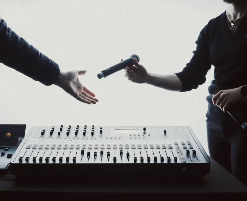 A microphone being passed between two people beside an audio mixing console.