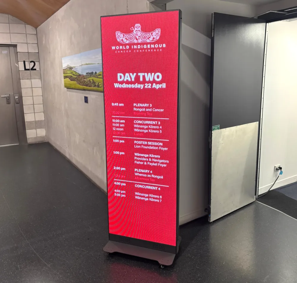 Freestanding digital poster displaying an event programme beside a lift.