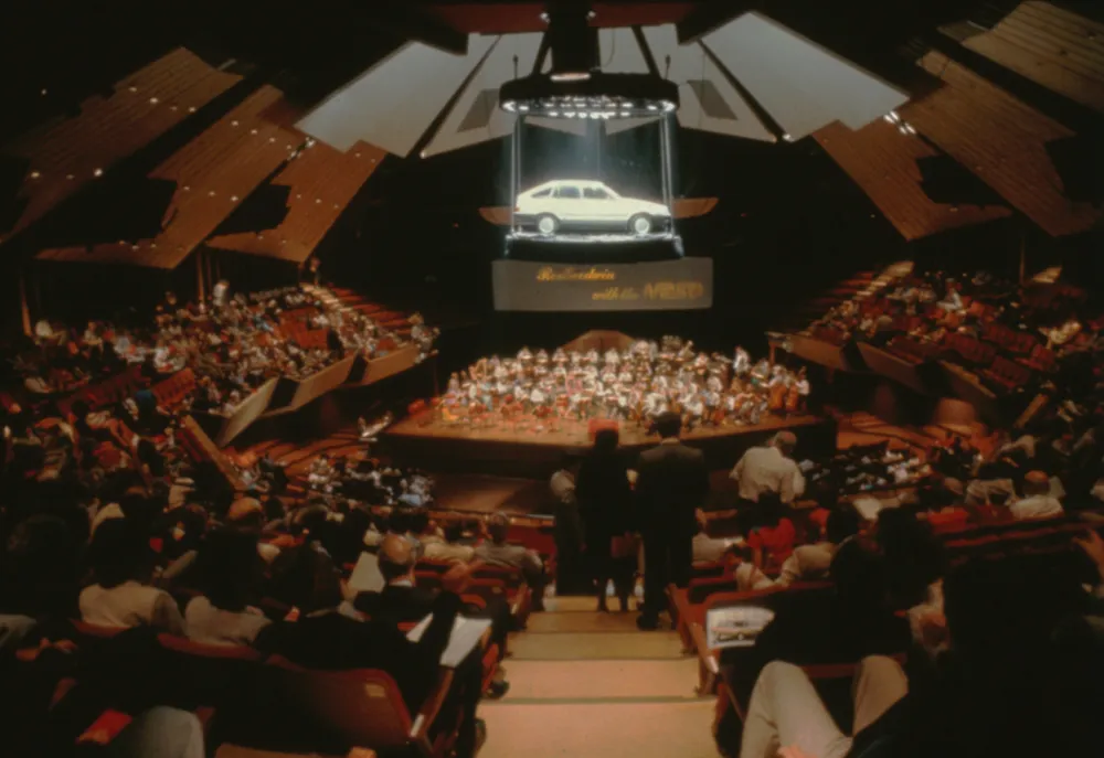 Archival photograph of a crowded event hall with a large presentation screen.